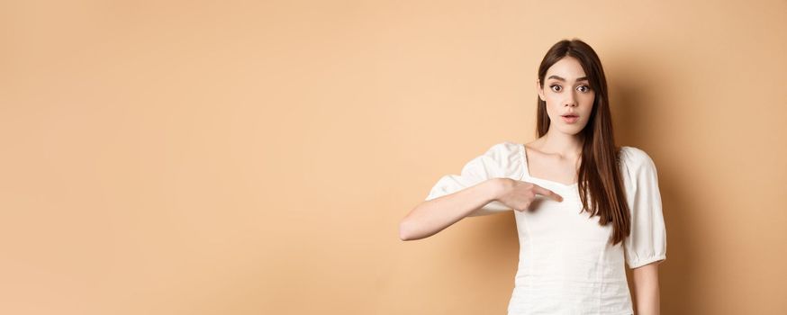 Confused girl pointing at herself with disbelief, being chosen or picked, standing on beige background.
