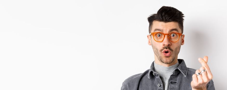 Close up portrait of cute guy with moustache wearing glasses, showing hand heart sign, look impressed at camera, standing on white background.