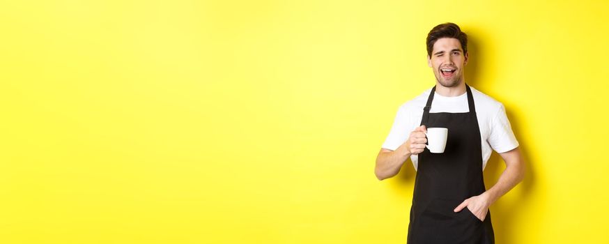 Cheerful barista in black apron serving coffee and winking, standing against yellow background.