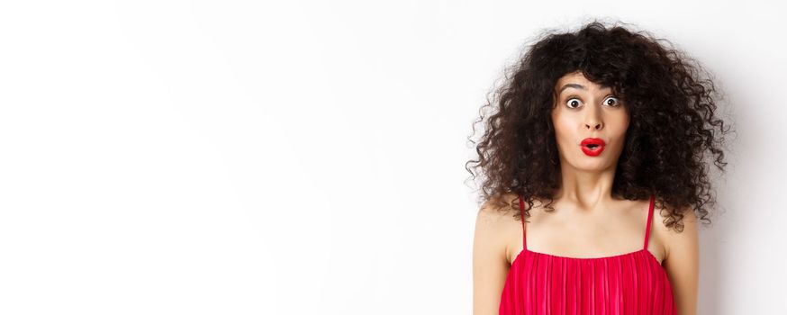 Close-up of surprised caucasian woman with red lips and curly hairstyle, saying wow and looking amazed, standing in red dress over white background.