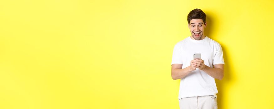 Surprised man reading text message on mobile phone, looking amazed and happy at smartphone screen, standing over yellow background.