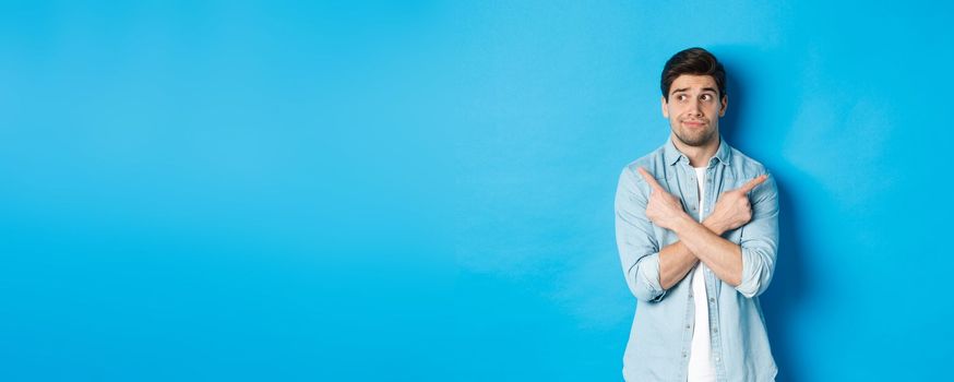 Portrait of indecisive adult man pointing fingers sideways but looking left, making choice between two products, standing against blue background.