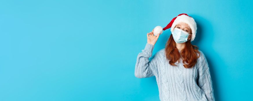 Christmas, quarantine and covid-19 concept. Cheerful teen redhead girl in santa hat and face mask, staring at camera pleased, standing confident against blue background.