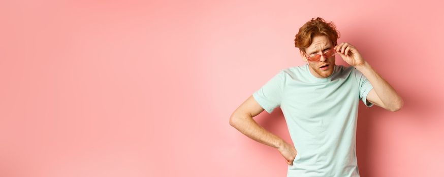 Tourism and vacation concept. Confused funny guy with red messy hair and beard, take-off glasses and frowning puzzled, standing over pink background.