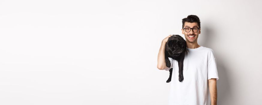 Cheerful hipster man in glasses, holding cute black pug dog on shoulder, staring at camera with happy smile, standing over white background.