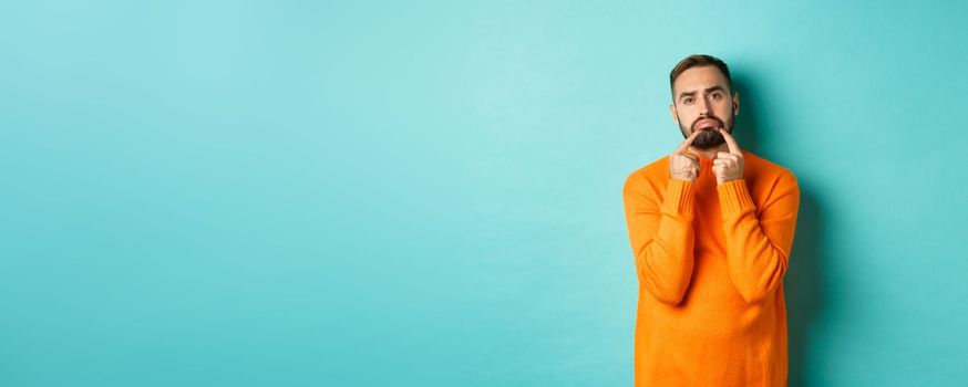 Image of gloomy bearded man, making sad face and frowning, standing upset in orange sweater against turquoise background.