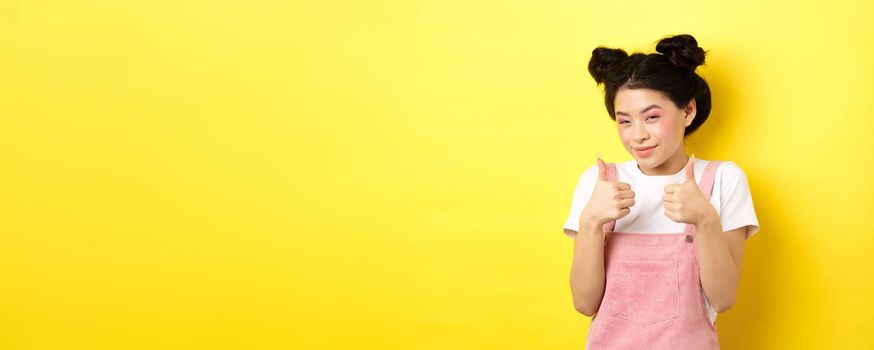 Cute teen asian girl with beauty makeup, showing thumbs up and smiling, praising and motivating you, making compliment, standing pleased on yellow background.