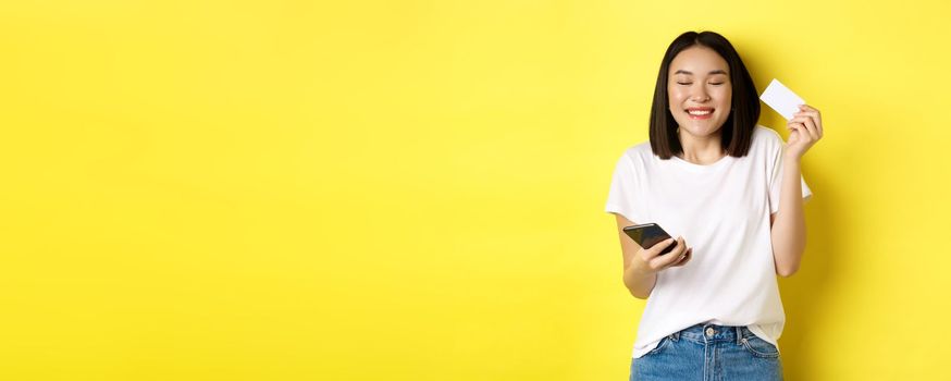 E-commerce and online shopping concept. Happy asian woman looking excited, buying something in internet, holding smartphone and showing plastic credit card, yellow background.