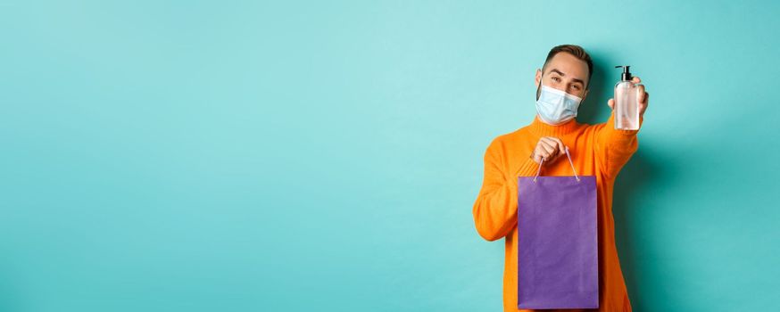 Coronavirus, pandemic and lifestyle concept. Man in face mask showing shopping bag and hand sanitizer, standing over turquoise background.