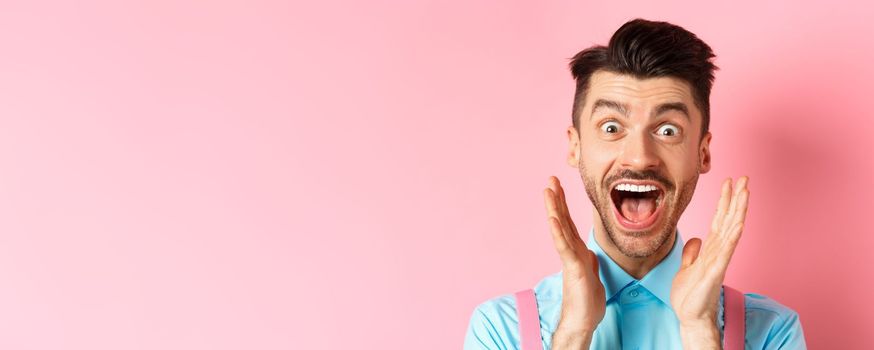 Close-up of happy caucasian guy screaming of joy and surprise, checking out advertisement, seeing something cool, standing over pink background.