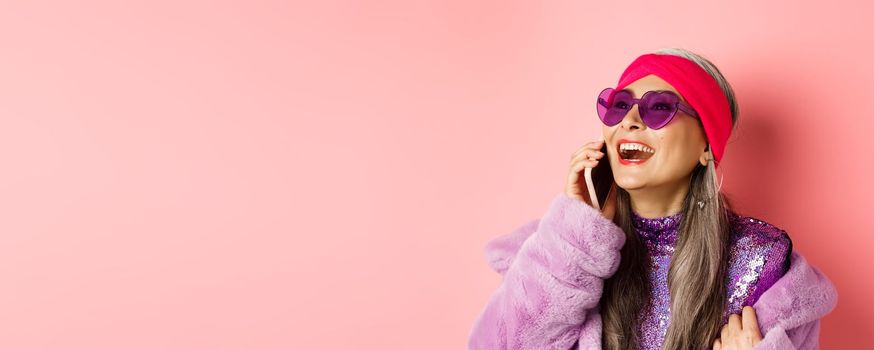 Fashion. Beautiful asian senior woman in trendy sunglasses and headband laughing, talking on mobile phone and looking aside, pink background.