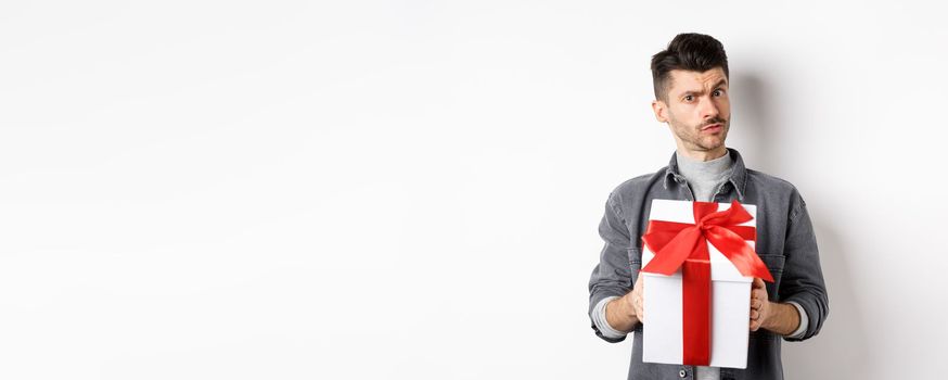 Surprised young man look with disbelief and hold surprise gift, raising eyebrow doubtful, being suspicious, standing on white background. Valentines day concept.