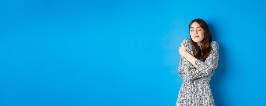 Romantic young natural woman in dress close eyes, smiling and hugging herself with love and tenderness, standing on blue background.