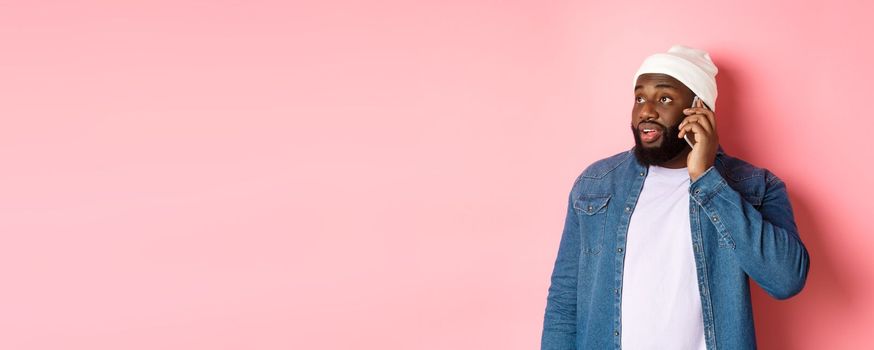 Hipster Black man talking on phone, looking left and having mobile conversation, standing over pink background.