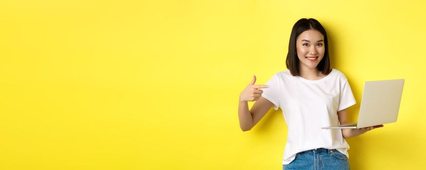 Smiling asian woman pointing finger at her laptop, showing something online, standing over yellow background.