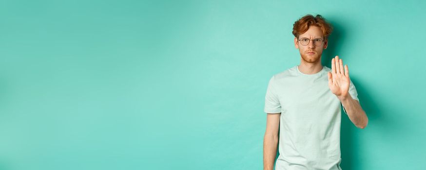 Angry and serious young man with red hair, wearing glasses, showing stop gesture, telling no, disapprove and prohibit something bad, standing over turquoise background.