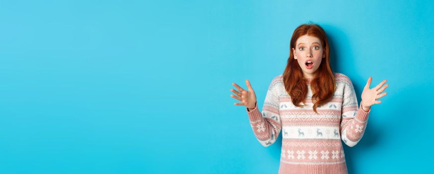 Image of startled redhead woman staring at camera with hands spread sideways, catching something, standing in winter sweater against blue background.