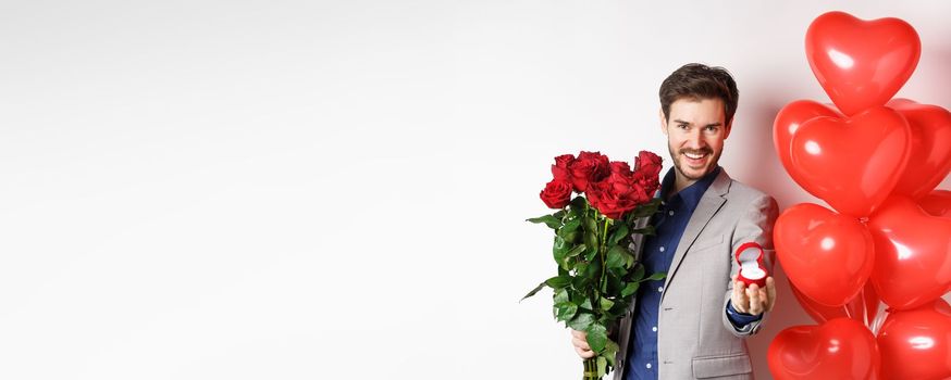 Handsome boyfriend in suit making a wedding proposal, showing engagement ring and say marry me, holding red roses, standing near Valentines day balloons, white background.