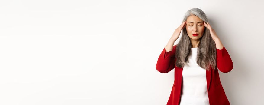 Distressed asian businesswoman trying to calm down, massaging temples on head to soothe headache, standing over white background.