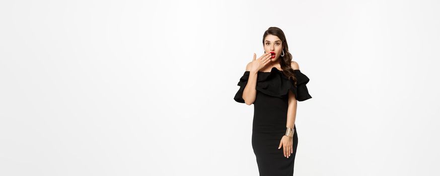 Beauty and fashion concept. Full length shot of beautiful and coquettish woman in black dress, open mouth and looking wondered at camera, standing over white background.