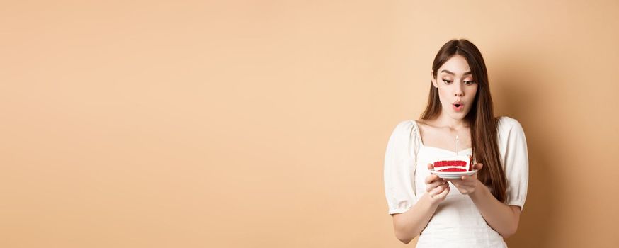 Beauty birthday girl blowing candle on b-day cake, making wish with excited face, standing on beige background and celebrating.