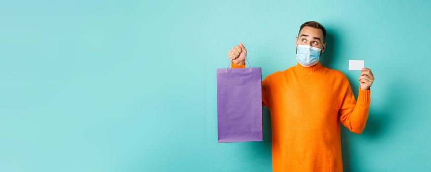 Covid-19, pandemic and lifestyle concept. Thoughtful man in face mask, holding purple shopping bag and credit card, thinking or imaging, standing over turquoise background.