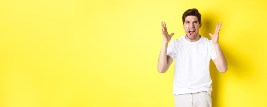 Angry man yelling and shaking hands, grimacing with hatred, standing hateful against yellow background. Copy space