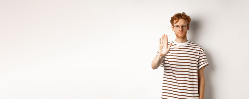 Serious young man with ginger hair frowning, showing stop gesture, refuse and prohibit something bad, standing displeased over white background.