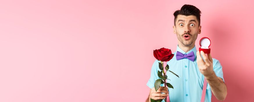 Valentines day. Funny man with moustache and bow-tie making proposal, showing engagement ring and propose with red rose, standing over pink background.