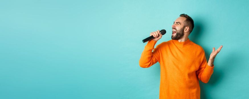 Handsome adult man perform song, singing into microphone, standing against turquoise background. Copy space