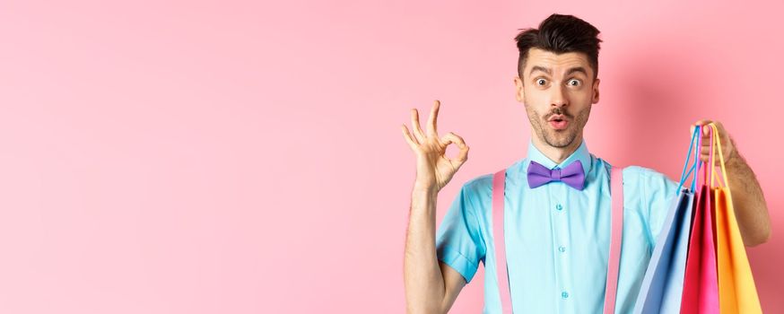 Funny guy in bow-tie showing OK sign and shopping bags, praising good deal in shops, standing over pink background.