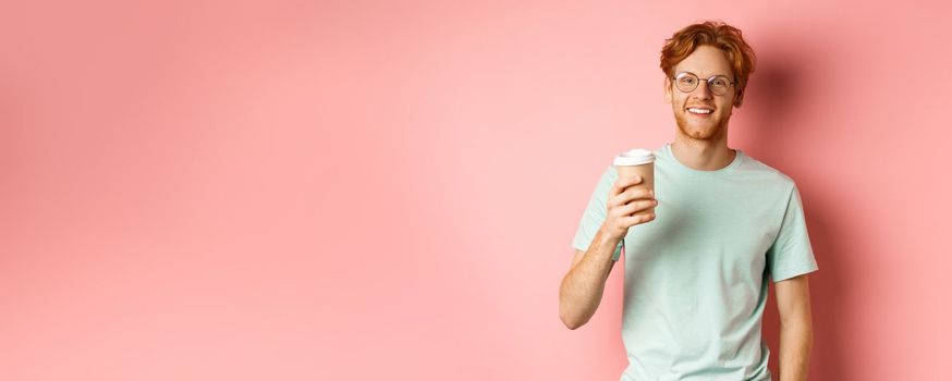 Happy redhead man in glasses and t-shirt drinking coffee and smiling, enjoying lunch break, holding takeaway cup, standing over pink background.