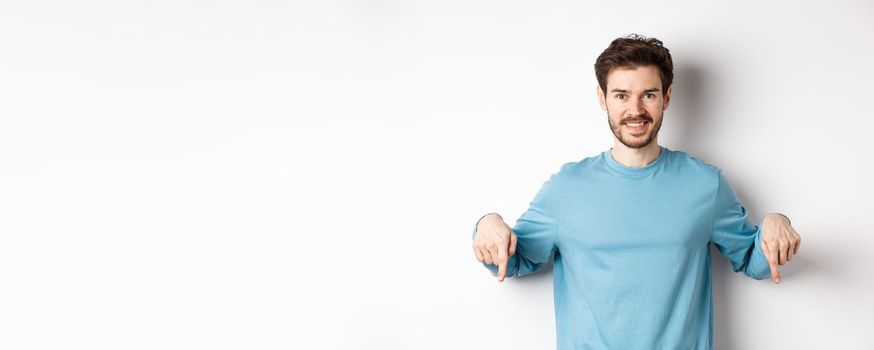 Smiling confident man pointing fingers down, showing promo banner or logo on white background, standing in casual blue sweatshirt.