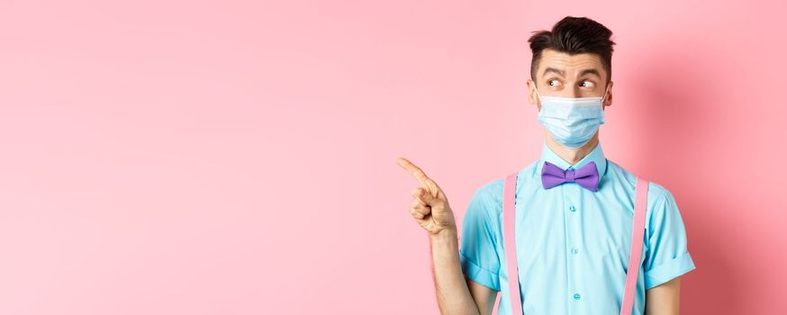 Covid-19, pandemic and health concept. Funny man in bow-tie and medical mask pointing finger left, looking aside curious, showing logo, standing on pink background.
