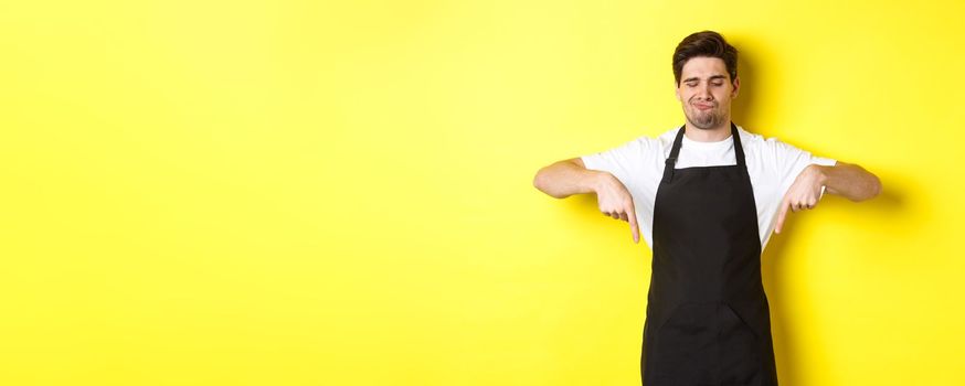 Doubtful barista complaining, pointing fingers down and grimacing displeased, standing in black apron against yellow background.