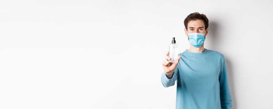 Covid-19, health and quarantine concept. Young man in medical mask stretch out hand and showing sanitizer, recommending antiseptic, standing over white background.