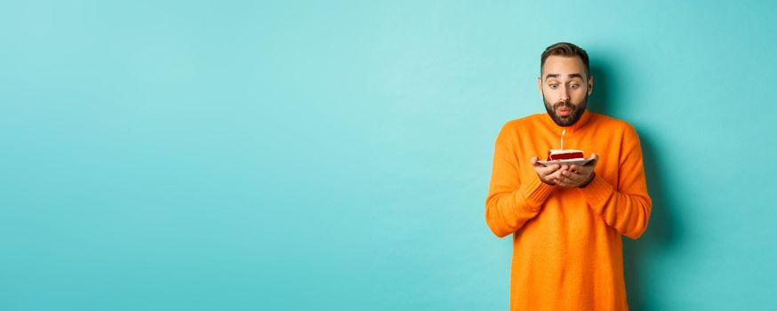 Handsome adult man celebrating birthday, blowing out candle on cake and making wish, standing against turquoise background.