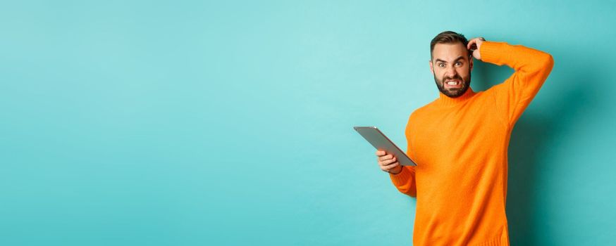 Confused man scratching head and looking worried, working on digital tablet, standing against turquoise background.