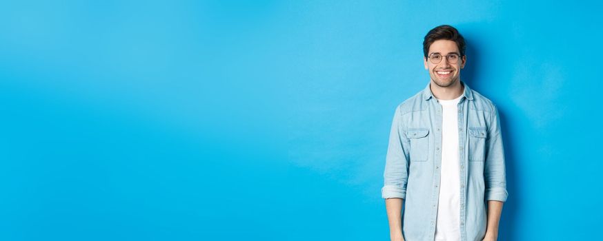 Young modern man in glasses and casual outfit standing against blue background, smiling happy at camera.