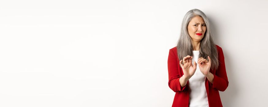 Disgusted asian businesswoman with grey hair, wearing red blazer and makeup, rejecting something disgusting, showing stop sign, white background.