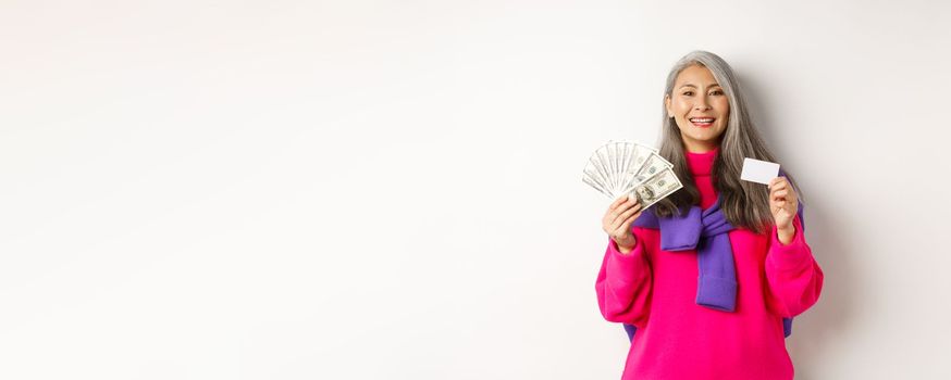 Shopping concept. Stylish asian senior woman with grey hair, showing money dollars and plastic credit card, standing over white background.