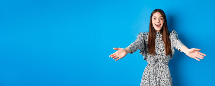 Beautiful happy woman in dress stretch out hands in warm welcome, smiling and greeting you, inviting guests, standing on blue background.