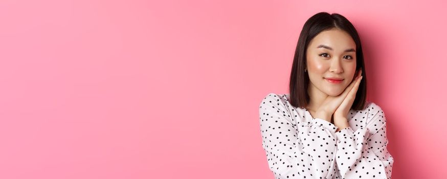 Beauty and lifestyle concept. Close-up of pretty korean girl leaning on palms, looking at camera with admiration, standing over pink background.