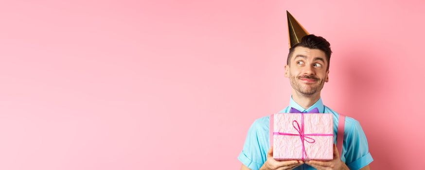 Holidays and celebration concept. Silly guy with moustache and bow-tie, wearing party hat, receive birthday gift and looking dreamy aside, standing over pink background.