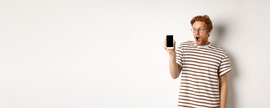 Technology and e-commerce concept. Surprised and shocked redhead guy checking out online promotion, showing blank smartphone screen and drop jaw, white background.