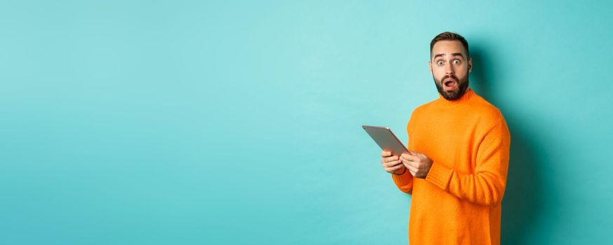Image of male model in orange sweater using digital tablet, looking surprised, standing over light blue background.