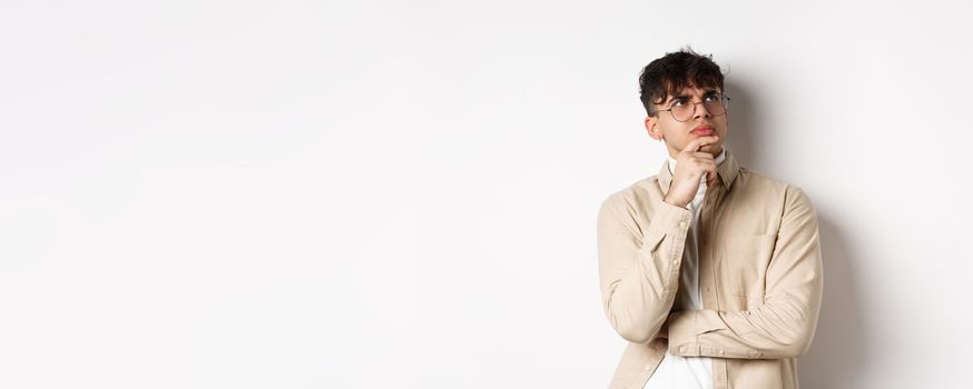 Handsome hipster guy in glasses look pensive at empty space, touching chin and frowning, stare up thoughtful, making choice with doubtful face, standing on white background.
