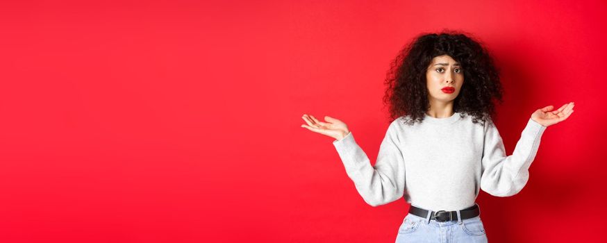 Who knows. Clueless young woman with curly hair, shrugging and looking confused, standing in casual clothes on red background.