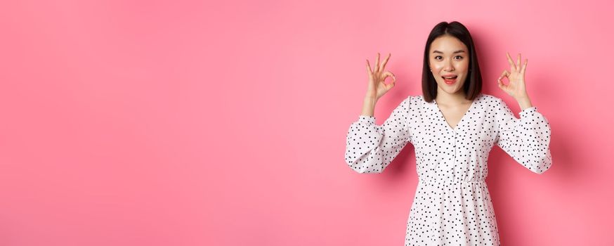 Pretty young asian woman in dress showing okay sign, praising and showing approval, looking satisfied, standing against pink background.