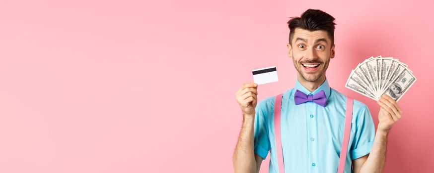 Cheerful caucasian man with moustache and bow-tie showing plastic credit card with money in dollars, smiling at camera, standing over pink background.